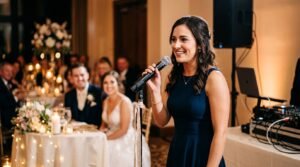 A speaker gives a wedding toast while newlyweds and guests blur in the background.