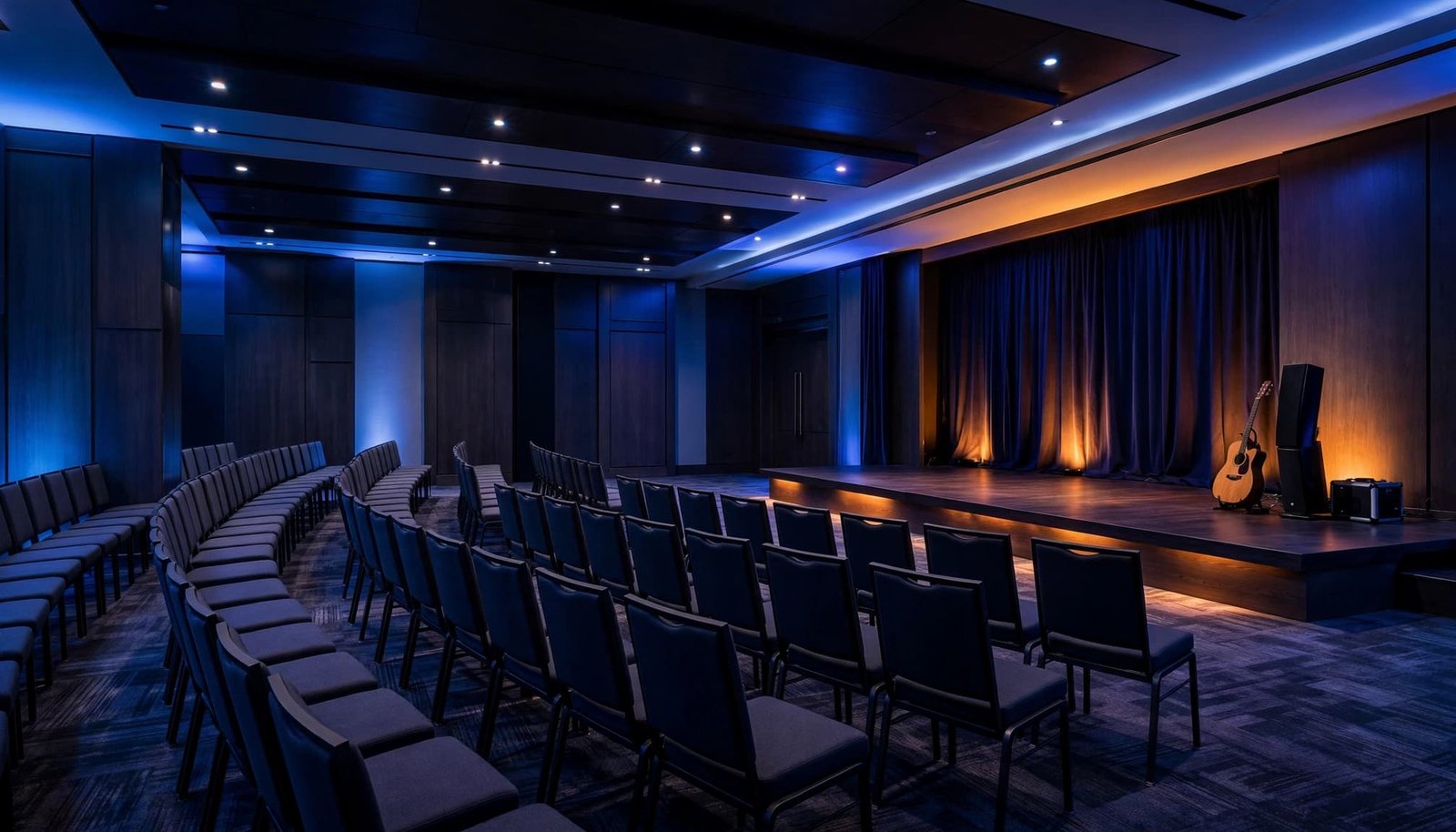 Empty conference room with stage, chairs, guitar, and speaker system under blue and amber lighting.